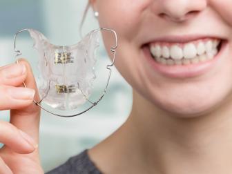 Eine junge Frau lächelt und hält eine durchsichtige lose Zahnspange in der Hand. Die Zahnspange ist gut sichtbar und wird vor dem Hintergrund einer Zahnarztpraxis präsentiert.