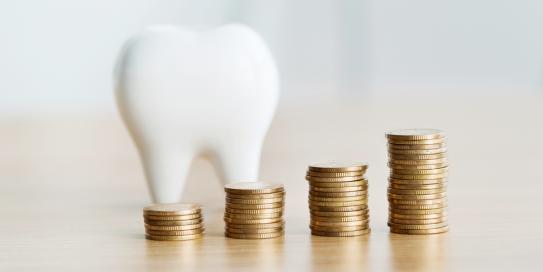 Symbolischer Zahn aus Keramik im Hintergrund, davor vier ansteigende Stapel aus Goldmünzen auf einem Holztisch – Darstellung steigender Zahnarztkosten oder Investitionen in Zahngesundheit.