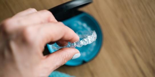 Hand einer Person, die eine transparente Zahnschiene (Aligner) aus einem blauen Aufbewahrungsbehälter nimmt – symbolisch für moderne kieferorthopädische Behandlung.
