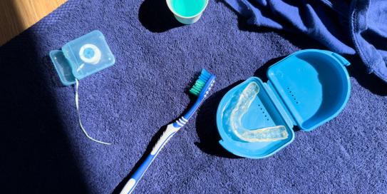Zahnpflegeset auf einem blauen Handtuch: Zahnbürste, Zahnseide, Mundspülung und transparente Zahnschiene in einem blauen Aufbewahrungsbehälter – dargestellt im Sonnenlicht.