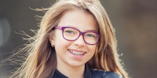 Fröhliches Mädchen mit langen Haaren und pinker Brille lächelt in die Kamera und zeigt ihre feste Zahnspange.