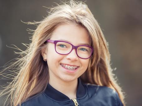 Fröhliches Mädchen mit langen Haaren und pinker Brille lächelt in die Kamera und zeigt ihre feste Zahnspange.