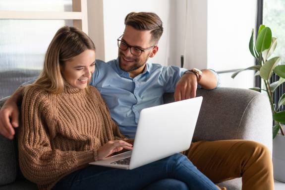 Junges Paar sitzt entspannt auf einem Sofa und schaut gemeinsam lachend auf einen Laptop – gemütliche Atmosphäre in einem modernen Wohnzimmer.