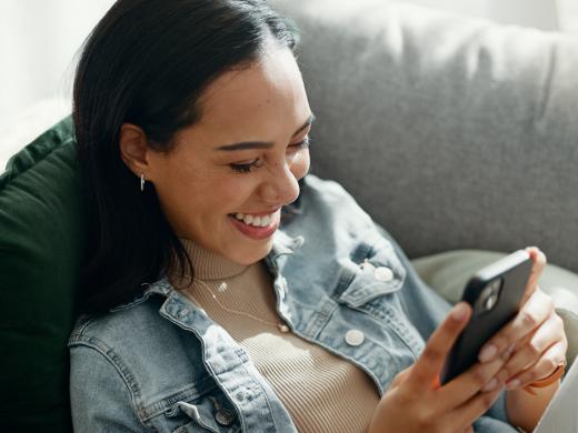 Junge Frau sitzt entspannt auf einem Sofa, lächelt und schaut auf ihr Smartphone – gemütliche und positive Stimmung.