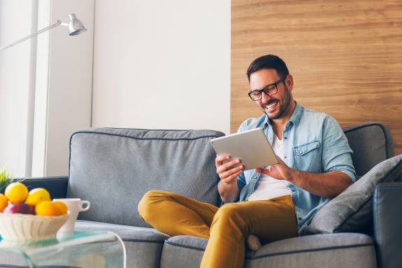 Lachender Mann sitzt entspannt auf einem grauen Sofa und schaut auf ein Tablet – moderne, gemütliche Wohnzimmeratmosphäre.