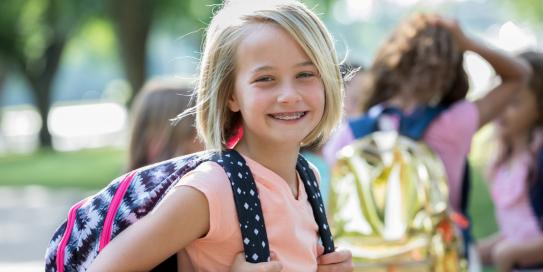 Lächelndes Mädchen mit Zahnspange und Rucksack auf dem Rücken steht draußen mit anderen Schulkindern – sonniger Tag im Park oder Schulhof.
