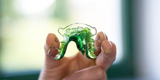 Hand hält eine grüne herausnehmbare Zahnspange vor einem unscharfen Fensterhintergrund