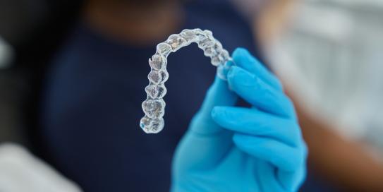 Zahnarzt oder Kieferorthopäde hält transparente Zahnschiene (Aligner) mit blauer Handschuhhand vor einem sitzenden Patienten.