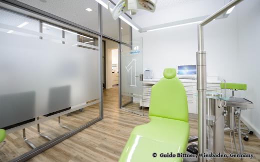 Moderner Behandlungsraum in einer Zahnarztpraxis mit grünem Behandlungsstuhl, Glaswänden und heller, freundlicher Einrichtung.