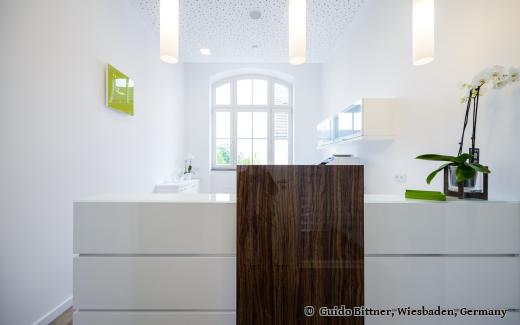 Moderne Rezeption einer Zahnarztpraxis mit weißen Möbeln, Holzakzenten und stilvoller Beleuchtung – im Hintergrund ein großes Fenster und eine Orchidee als Dekoration.