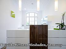 Moderne Rezeption einer Zahnarztpraxis mit weißen Möbeln, Holzakzenten und stilvoller Beleuchtung – im Hintergrund ein großes Fenster und eine Orchidee als Dekoration.