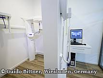 Röntgenraum in einer Zahnarztpraxis mit digitalem Panorama-Röntgengerät und einem Monitor, auf dem ein Kieferbild angezeigt wird; moderne, saubere Einrichtung mit Holzfußboden.