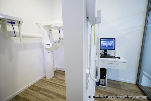 Röntgenraum in einer Zahnarztpraxis mit digitalem Panorama-Röntgengerät und einem Monitor, auf dem ein Kieferbild angezeigt wird; moderne, saubere Einrichtung mit Holzfußboden.