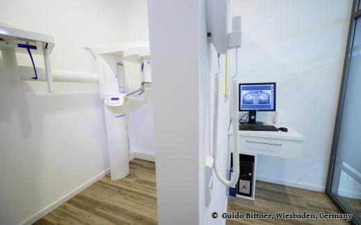 Röntgenraum in einer Zahnarztpraxis mit digitalem Panorama-Röntgengerät und einem Monitor, auf dem ein Kieferbild angezeigt wird; moderne, saubere Einrichtung mit Holzfußboden.