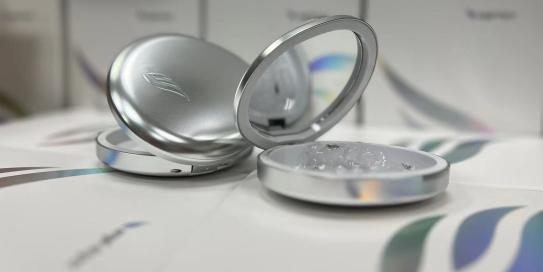 Silberfarbene Dose mit Spiegeldeckel, geöffnet und mit einer transparenten Substanz gefüllt, steht auf einer Oberfläche mit modern gestaltetem Verpackungsdesign im Hintergrund. Die Szene wirkt hochwertig und kosmetisch orientiert.